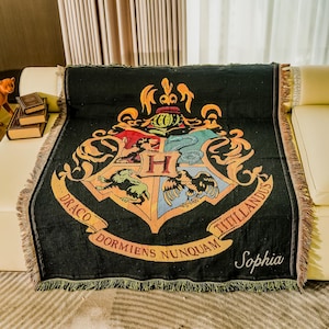 May include: Black woven throw blanket featuring a colorful crest design with the words "Draco Dormiens Nunquam Titillandus" and the name "Sophia". The blanket has a fringed edge and is draped over a cream-colored sofa.