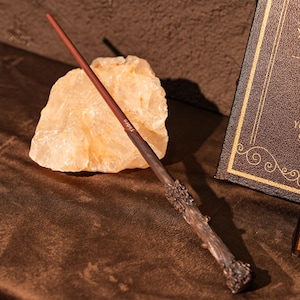 May include: A brown wand with a textured handle and a reddish-brown shaft. The word "Jaden" is printed on the wand. The wand is resting on a brown surface next to a light-coloured rock and a book.