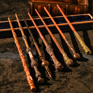 May include: A collection of seven intricately designed wands, each with unique features. The wands vary in color, including shades of brown, tan, and green. Some wands have detailed handles with ornate carvings, while others have smooth, sleek designs. The wands are arranged on a dark surface.