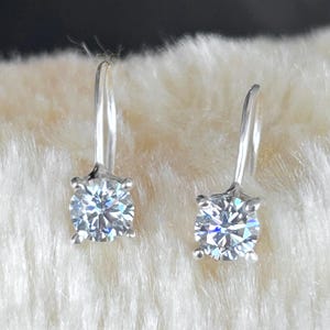 Può includere: Un paio di orecchini pendenti in argento con pietre preziose rotonde e trasparenti. Le pietre sono incastonate in una montatura a quattro griffe e riflettono la luce, creando un effetto scintillante. Gli orecchini sono esposti su una superficie bianca e soffice.