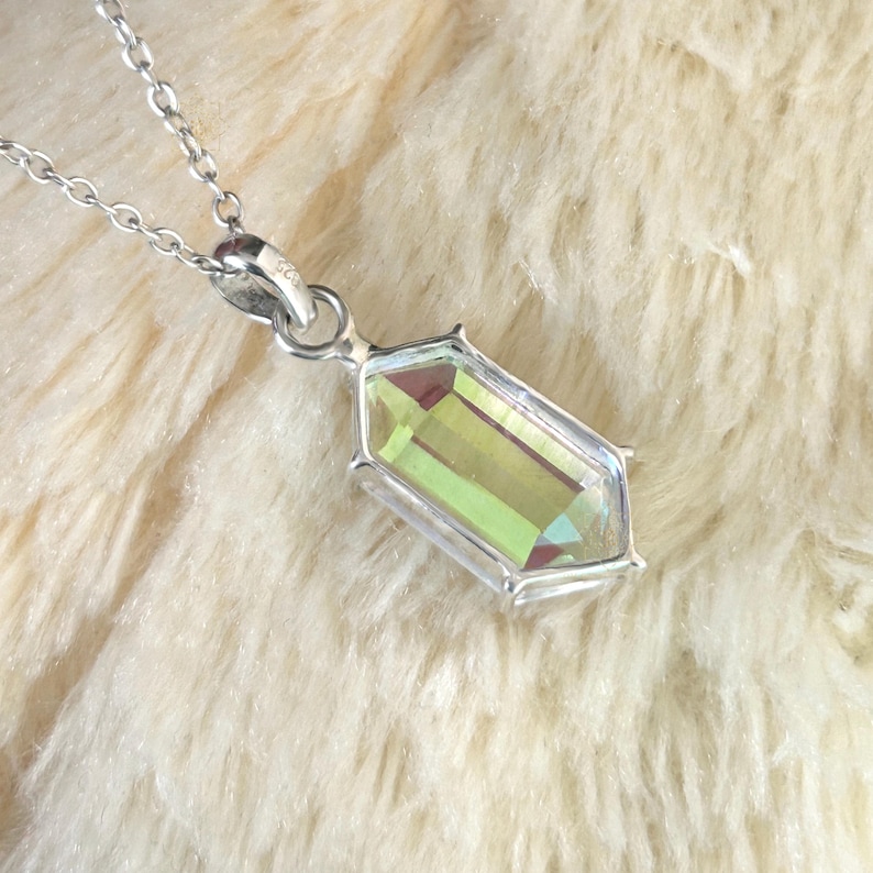 May include: A silver necklace with a rectangular pendant. The pendant features a large, iridescent, emerald-cut gemstone with a silver bezel. The chain is delicate and the pendant is centered in the frame. The background is a soft, cream-colored, fuzzy texture.