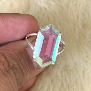 Pu&ograve; includere: Un anello d'argento con una grande gemma rosa rettangolare con qualit&agrave; iridescenti. La gemma &egrave; incastonata in una fascia d'argento dal design semplice ed elegante. L'anello &egrave; tenuto da una mano su uno sfondo morbido e chiaro.
