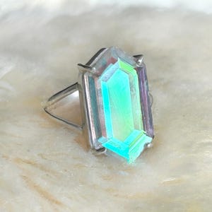 Pu&ograve; includere: Un anello d'argento con una grande pietra preziosa iridescente rettangolare. La pietra mostra uno spettro di colori, tra cui verde, blu e viola, incastonata in una fascia d'argento. L'anello &egrave; esposto su una superficie bianca e morbida.