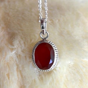 Peut inclure: Un collier pendentif en argent avec une pierre pr&eacute;cieuse ovale, facett&eacute;e, rouge fonc&eacute;. La pierre est sertie dans une lunette en argent avec une bordure en forme de corde et est suspendue &agrave; une fine cha&icirc;ne en argent. Le fond est un tissu doux de couleur cr&egrave;me.