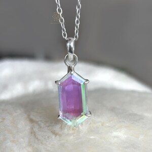 May include: A silver necklace featuring a pendant with a rectangular, iridescent crystal. The crystal displays shades of purple, pink, and green. The pendant is set in a silver frame and hangs from a delicate silver chain. The jewelry is displayed on a soft, white surface.