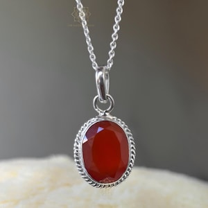 Peut inclure: Un collier pendentif en argent avec une pierre pr&eacute;cieuse rouge ovale et facett&eacute;e. La pierre est sertie dans un cadre argent&eacute; avec un motif de corde torsad&eacute;e. Le collier a une cha&icirc;ne en argent d&eacute;licate.