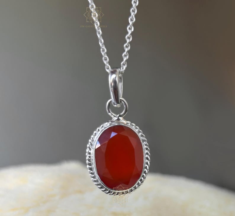Peut inclure: Un collier pendentif en argent avec une pierre pr&eacute;cieuse rouge ovale et facett&eacute;e. La pierre est sertie dans un cadre argent&eacute; avec un motif de corde torsad&eacute;e. Le collier a une cha&icirc;ne en argent d&eacute;licate.