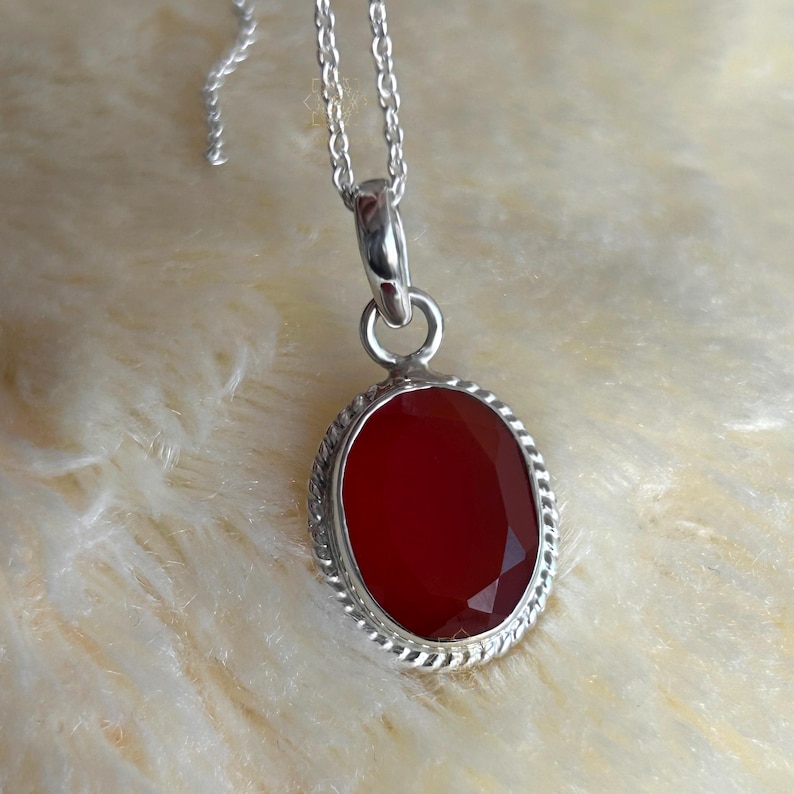 Peut inclure: Un collier pendentif en argent avec une pierre pr&eacute;cieuse rouge fonc&eacute; ovale et facett&eacute;e. La pierre est sertie dans un cadre argent&eacute; avec un motif de corde torsad&eacute;e. La cha&icirc;ne est fine, id&eacute;ale pour un usage quotidien.