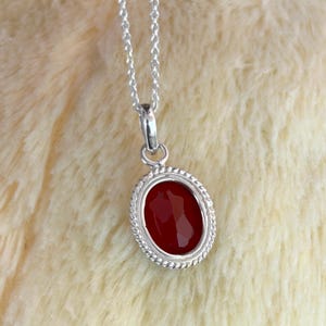 Peut inclure: Un collier en argent avec un pendentif ovale en pierre rouge facett&eacute;e. Le pendentif est encadr&eacute; d'un motif torsad&eacute;. La cha&icirc;ne du collier est fine et argent&eacute;e. Le fond est une surface textur&eacute;e douce et de couleur claire.