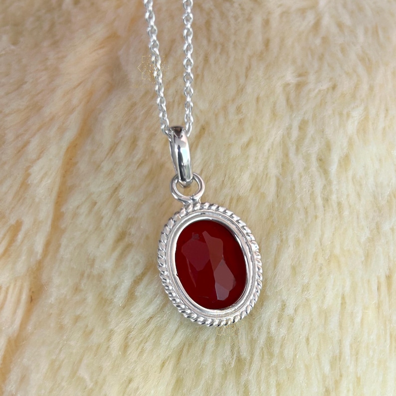 Peut inclure: Un collier en argent avec un pendentif ovale en pierre rouge facett&eacute;e. Le pendentif est encadr&eacute; d'un motif torsad&eacute;. La cha&icirc;ne du collier est fine et argent&eacute;e. Le fond est une surface textur&eacute;e douce et de couleur claire.