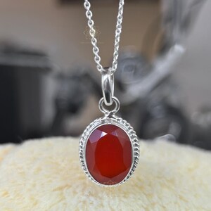 Peut inclure: Un collier en argent avec un pendentif ovale en pierre rouge facett&eacute;e. Le pendentif est serti dans un cadre argent&eacute; avec un motif de corde torsad&eacute;e. La cha&icirc;ne est d&eacute;licate et suspendue &agrave; une petite boucle.