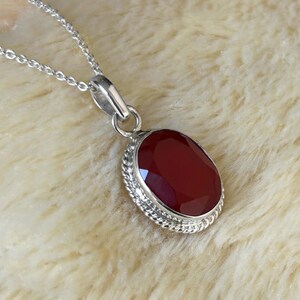 Peut inclure: Un collier pendentif en argent avec une pierre pr&eacute;cieuse rouge fonc&eacute; ovale et facett&eacute;e. La pierre est sertie dans une lunette en argent avec un motif de corde torsad&eacute;e. La cha&icirc;ne du collier est une fine cha&icirc;ne en argent.
