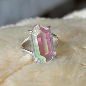 Pu&ograve; includere: Un anello d'argento con una grande gemma rettangolare dai colori iridescenti rosa, verde e giallo. La gemma sfaccettata cattura la luce creando un effetto scintillante. Il gambo dell'anello si divide in due fili su entrambi i lati della gemma.