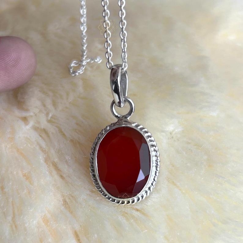 Peut inclure: Collier pendentif en argent avec une pierre pr&eacute;cieuse rouge ovale et facett&eacute;e. La pierre est sertie dans un cadre argent&eacute; avec un motif de corde torsad&eacute;e. La cha&icirc;ne du collier est &eacute;galement argent&eacute;e et d&eacute;licate.
