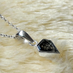 May include: A silver necklace featuring a geometric pendant with a black and clear crystal. The pendant has a unique shape and is suspended from a delicate silver chain. The crystal has black inclusions.
