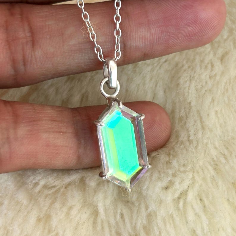 May include: A silver necklace featuring a rectangular pendant with iridescent green and yellow hues. The pendant is encased in a silver frame and hangs from a delicate silver chain. The necklace is displayed against a soft, off-white background.