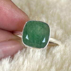 May include: A silver ring featuring a square-cut, faceted green gemstone with rounded corners. The stone displays a deep, mottled emerald green hue with visible internal texture, set within a smooth, polished silver bezel on a thin band.