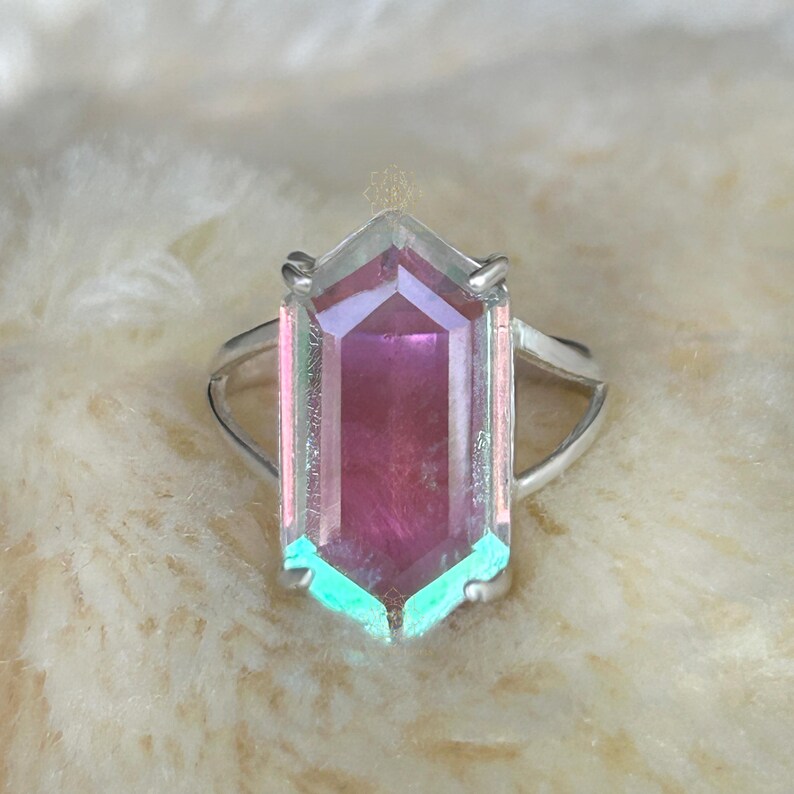 Pu&ograve; includere: Anello d'argento con una grande pietra preziosa iridescente a forma esagonale. La gemma presenta sfumature di rosa, viola e verde acqua, con una superficie trasparente e sfaccettata. La fascia dell'anello &egrave; semplice e sottile.