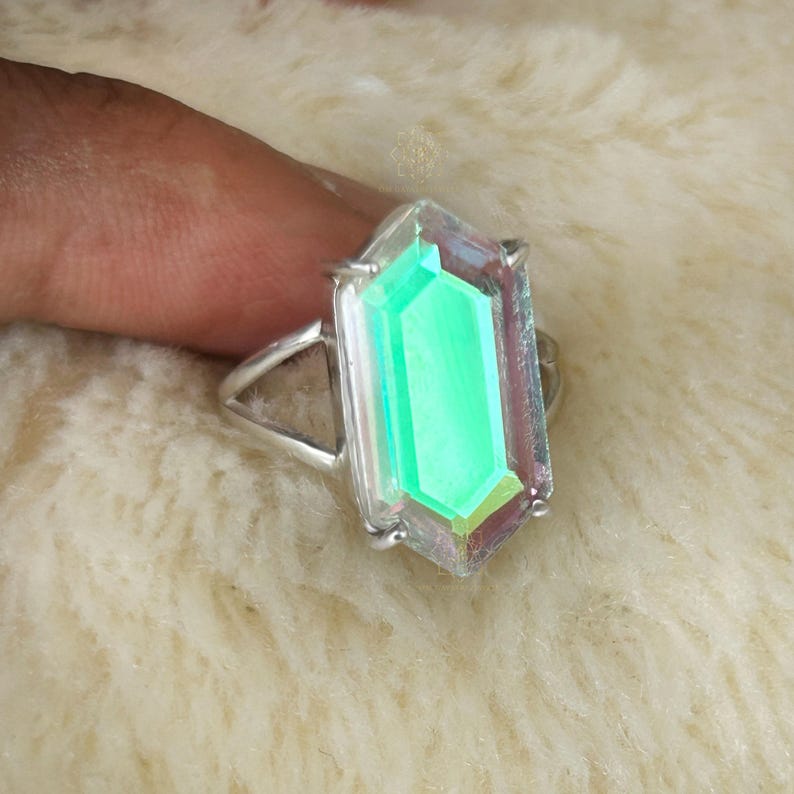 Pu&ograve; includere: Anello in argento con una grande gemma rettangolare dai colori verdi e iridescenti. La gemma &egrave; incastonata in una fascia d'argento, creando un design moderno. L'anello &egrave; mostrato su una superficie bianca e morbida.