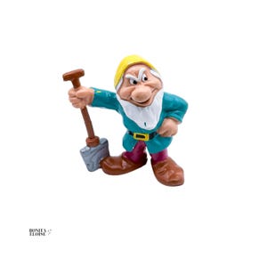 Pode incluir: Uma estatueta do Zangado da Branca de Neve, segurando uma pá. A figura está vestida com uma túnica turquesa, calças bordô e botas marrons. Ele usa um chapéu amarelo, tem barba branca e uma expressão rabugenta.