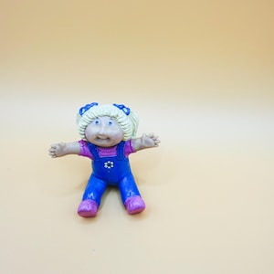 May include: A Cabbage Patch Kids doll figurine. The doll has blonde hair with blue bows, blue eyes, and is wearing a purple shirt and blue dungarees with a flower button. The doll's shoes are pink.