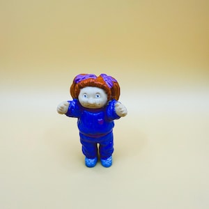 Op de afbeelding: Een vintage Cabbage Patch Kids poppenbeeldje. De pop heeft een blauwe outfit met een rood hart, blauwe schoenen en oranje en paars haar. De armen van de pop zijn uitgestrekt.