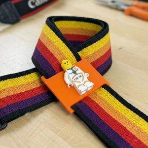 May include: A colorful camera strap with a rainbow stripe design, featuring a small Lego astronaut figure on an orange plastic piece. The strap is black on the edges and has a Canon logo on the end.