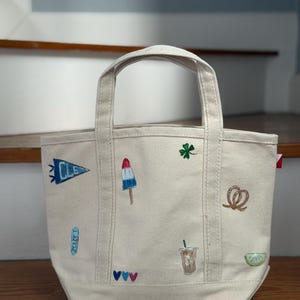 May include: A cream-colored canvas tote bag featuring small embroidered designs including a blue pennant with the text CLE, a red, white, and blue popsicle, a green four-leaf clover, a brown pretzel, a coffee cup, and a lime wedge.