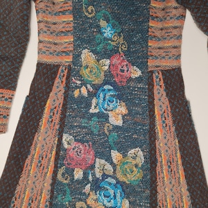 May include: A long-sleeved, knitted dress featuring a floral design. The dress has a teal center panel with embroidered roses in shades of pink, blue, and yellow. The sleeves and side panels are brown with orange and tan accents.
