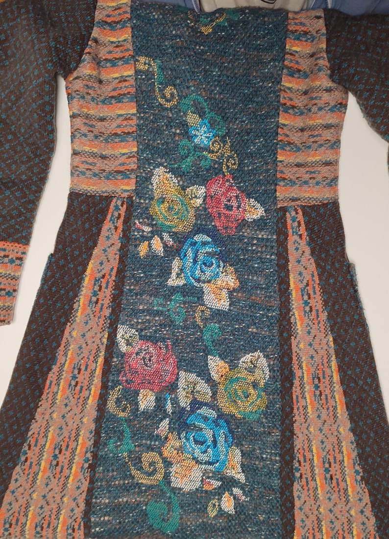 May include: A long-sleeved, knitted dress featuring a floral design. The dress has a teal center panel with embroidered roses in shades of pink, blue, and yellow. The sleeves and side panels are brown with orange and tan accents.