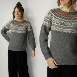 May include: A gray sweater with a patterned yoke featuring white, orange, and brown geometric designs. The long-sleeved sweater has a crew neck and is made of a soft, possibly wool, material. The sweater is paired with black pants.
