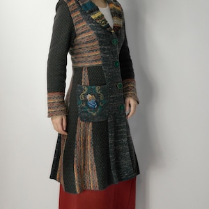 May include: A long, patchwork coat in shades of gray, orange, and blue. The coat features a patterned collar, sleeves, and pocket, with green buttons. The coat is knee-length and has a flared skirt. The coat is worn over a red skirt.