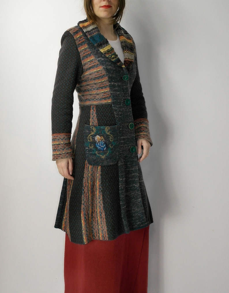 May include: A long, patchwork coat in shades of gray, orange, and blue. The coat features a patterned collar, sleeves, and pocket, with green buttons. The coat is knee-length and has a flared skirt. The coat is worn over a red skirt.