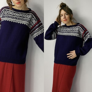 Puede incluir: Un suéter azul marino con un patrón geométrico blanco y rojo en el pecho y las mangas. El suéter tiene cuello redondo y mangas largas. La parte inferior del suéter es de color azul marino liso.