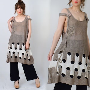 May include: A sleeveless taupe knit tunic featuring a scoop neckline, shoulder straps with circular accents, and a decorative spiral detail. The lower section displays a white and taupe cutout pattern with circular openings over a black base layer.
