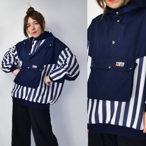 May include: A navy blue and white striped pullover jacket with a hood and a large front pocket. The jacket features a navy blue upper section and sleeves with white and navy stripes. The brand name "rukka" is visible on the pocket.