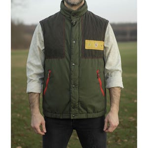 Può includere: Gilet trapuntato verde oliva e marrone con collo alto e cerniere rosse. Il gilet presenta una toppa gialla con un emblema a forma di diamante. Il gilet è abbottonato e presenta un design trapuntato.
