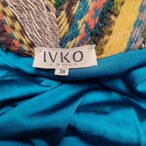 May include: Close-up of a garment label with the brand name "IVKO WOMAN" and size "38". The label is set against a teal fabric. The garment's collar features a colorful woven pattern with yellow, blue, orange, and green hues.