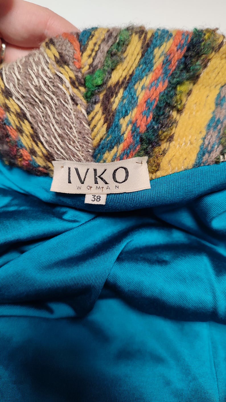 May include: Close-up of a garment label with the brand name "IVKO WOMAN" and size "38". The label is set against a teal fabric. The garment's collar features a colorful woven pattern with yellow, blue, orange, and green hues.