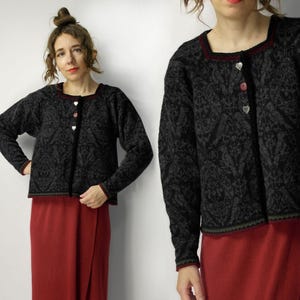 Peut inclure: Cardigan noir à motifs avec encolure carrée et manches longues. Le cardigan a des boutons en forme de cœur et une bordure rouge. La modèle porte une jupe rouge. Le motif est de couleur grise.
