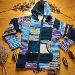 Puede incluir: Una sudadera con capucha de punto patchwork en tonos azules, morados y negros. La chaqueta tiene capucha, cremallera frontal y bolsillo de canguro. Las mangas y el cuerpo están hechos de varios cuadrados de punto. La chaqueta se muestra sobre una superficie de madera.