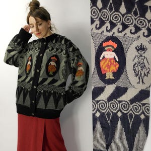 Op de afbeelding: Een vintage zwarte, groene en grijze cardigan met een knoopsluiting. De trui heeft geborduurde figuren in ovale kaders en geometrische patronen. De manchetten en kraag zijn zwart.