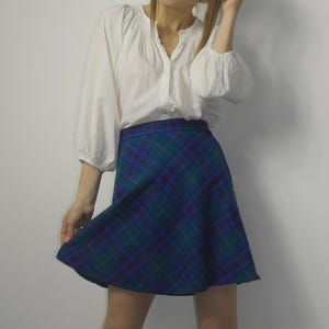 May include: A white blouse with a gathered neckline and long sleeves is worn with a plaid skirt. The skirt has a green and blue tartan pattern. The a-line skirt falls above the knees.