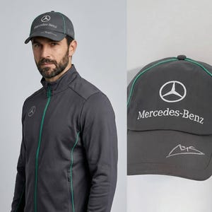 Può includere: Un cappellino da baseball grigio antracite con finiture verdi e il logo Mercedes-Benz in bianco. Il cappellino ha una visiera curva e una firma. La giacca grigia abbinata ha finiture verdi e il logo Mercedes-Benz.