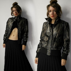 Puede incluir: Chaqueta bomber negra y dorada texturizada con cuello alto y cierre de botones. La chaqueta tiene dobladillo y puños de canalé. La modelo lleva un top negro y una falda larga plisada negra.