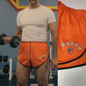May include: Bright orange athletic shorts with black edging and the white "KARHU" logo. These lightweight shorts are suitable for running or other sports. The image shows the shorts in a gym environment.