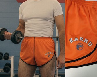 Può includere: Pantaloncini sportivi arancioni con bordi neri e logo "KARHU" bianco. Questi pantaloncini leggeri sono adatti per la corsa o altri sport. L'immagine mostra i pantaloncini in un ambiente di palestra.