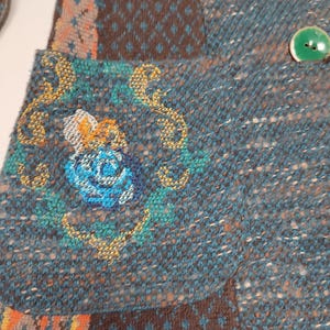 May include: Close-up of a teal and brown patchwork jacket with green buttons. The jacket features a floral embroidery on the pocket and a mix of patterned fabrics. The overall style is bohemian and handcrafted.