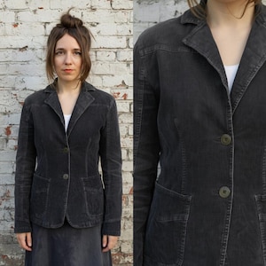 May include: A charcoal grey blazer with two front pockets and a single-button fastening. The blazer features a notched lapel and long sleeves. The person in the image is wearing a white top. The background is a white brick wall.