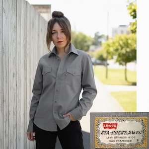 Peut inclure: Une chemise grise à manches longues avec deux poches de poitrine et une fermeture boutonnée. La chemise a une petite étiquette rouge Levi's sur une poche. L'étiquette Levi's Sta-Prest est visible. La chemise est de taille S.